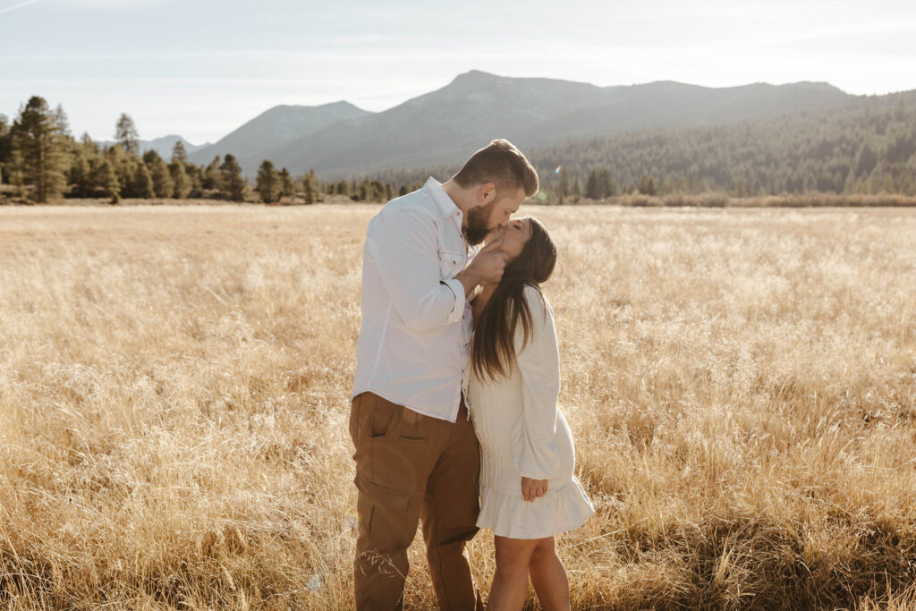 Couple kissing while man holds girlfriends face and standing together in golden grass field in Lake Tahoe with trees and mountains in background