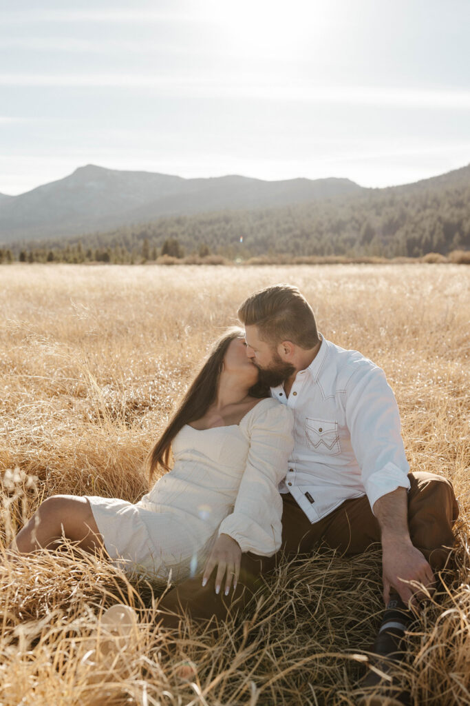 Couple sitting together in dry grass field while kissing in Lake Tahoe with trees and mountains in background