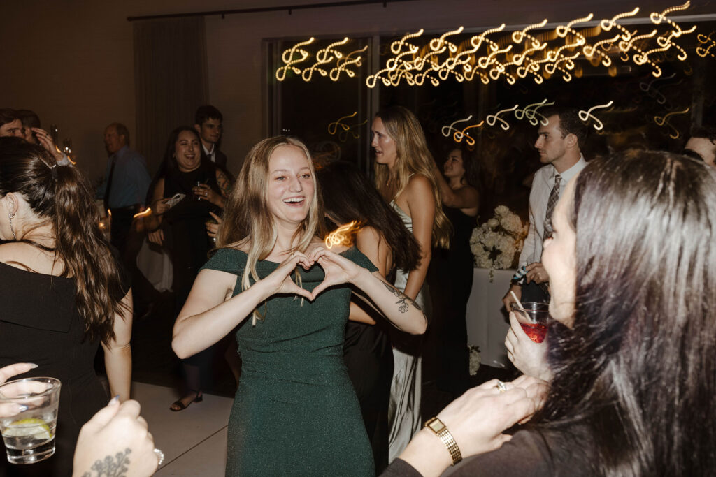 Wedding guests making hearts with hands at each other during reception  while other guests celebrate in background with string lights during reception in San Diego