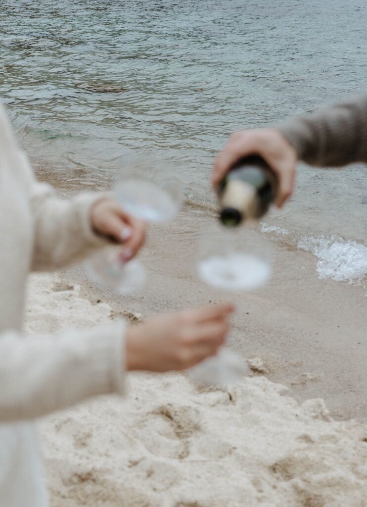 Engagement couple pouring champagne into glasses while on a beach in Lake Tahoe