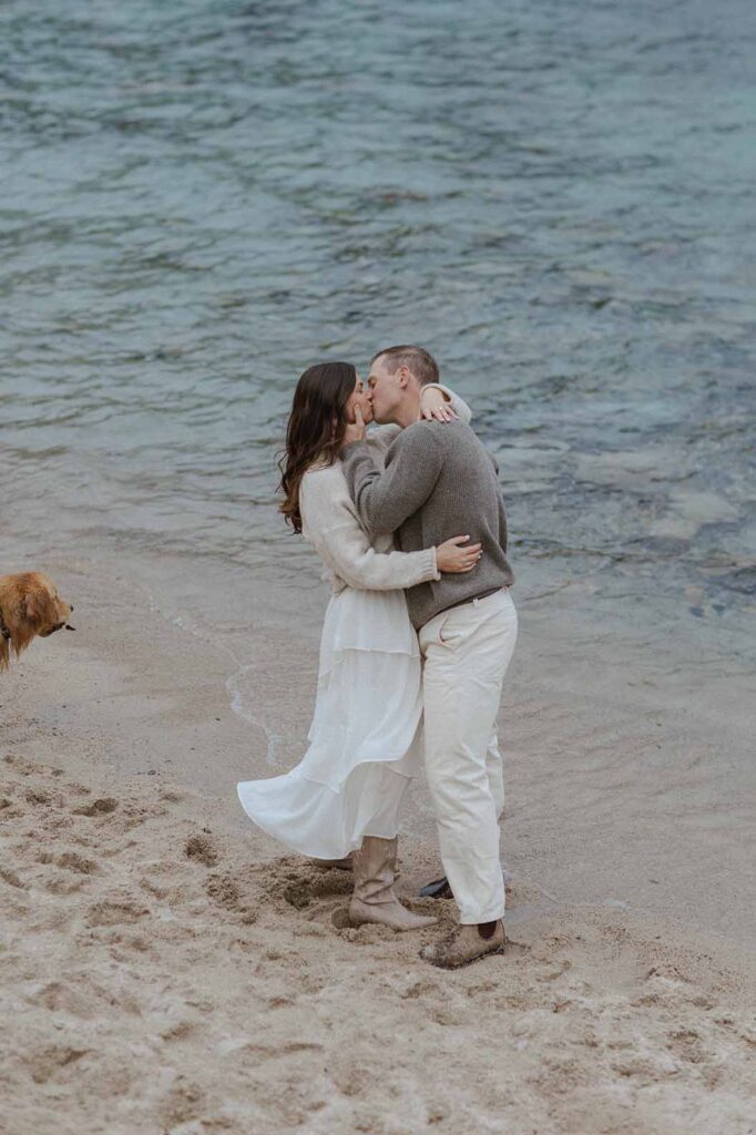 Engagement couple hugging each other while kissing on sandy beach in Lake Tahoe with water in background
