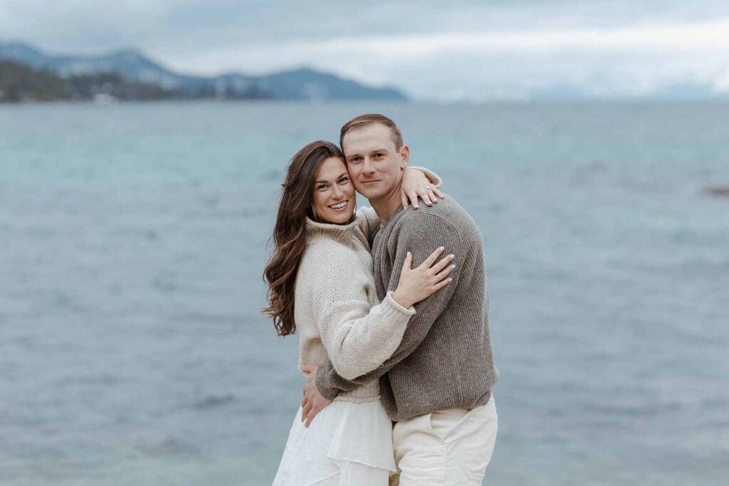 Engagement couple hugging while looking at the camera with Lake Tahoe in background