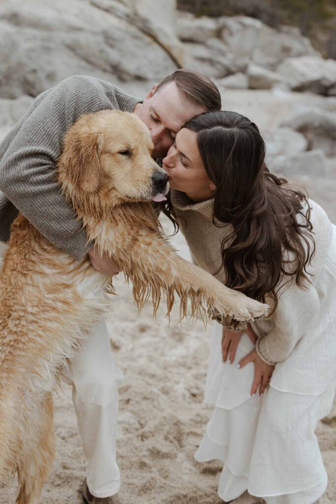 Engagement couple holding dog while kissing cheek on a sandy beach in Lake Tahoe