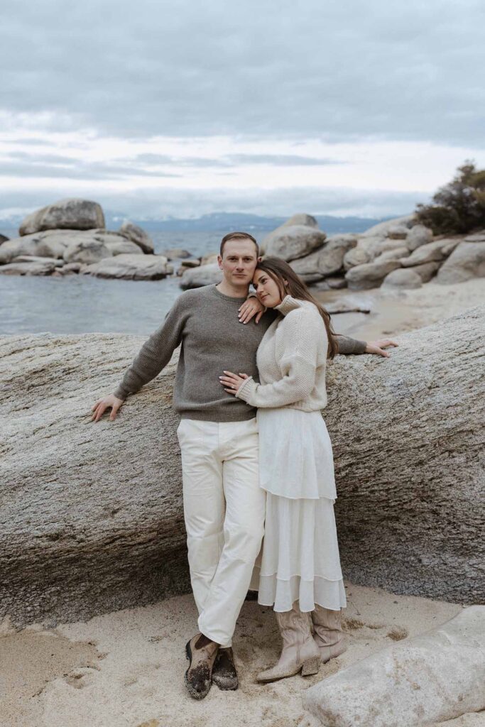 Engagement couple leaning on boulder together on a sandy beach in Lake Tahoe with storm clouds in background