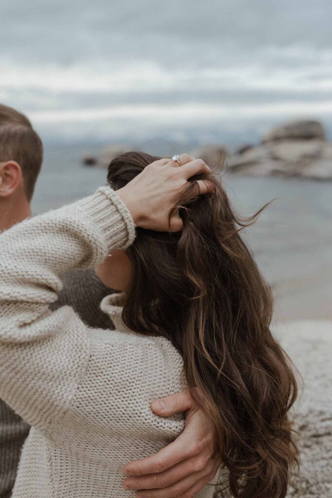 Woman playing with hair while wearing engagement ring and fiancé has arm around her in Lake Tahoe