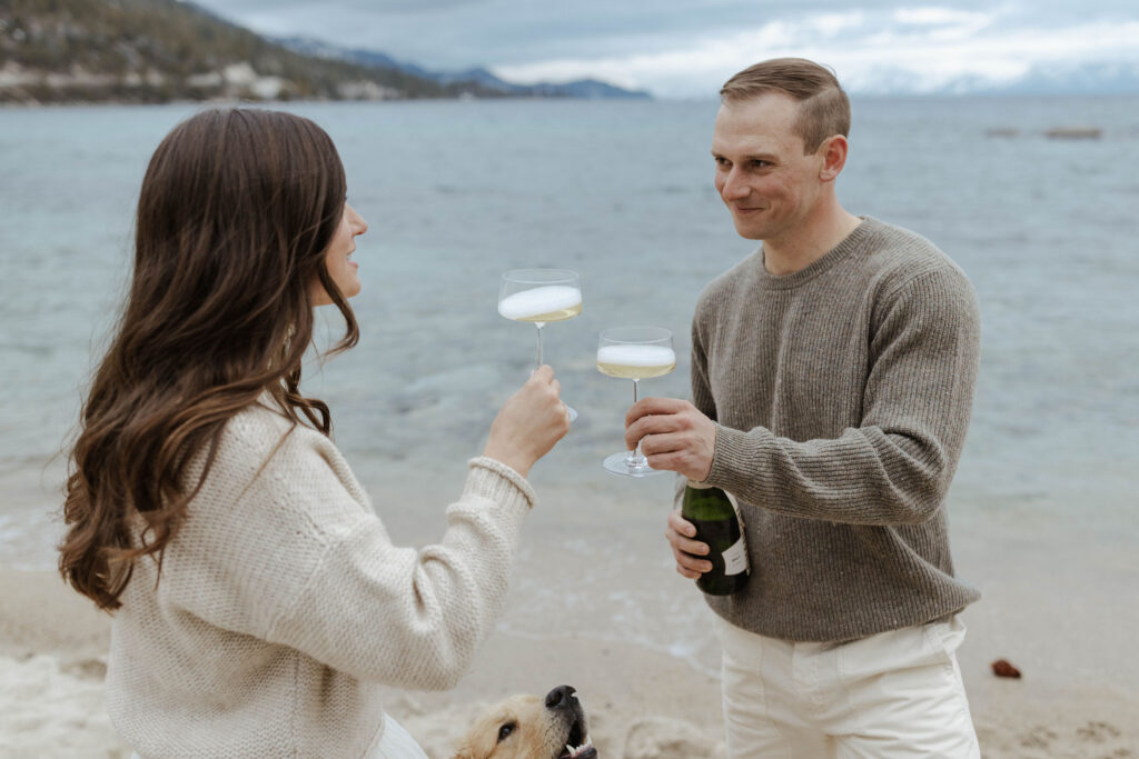 Man smiling at fiancé while cheering champagne glasses together on a sandy beach in Lake Tahoe