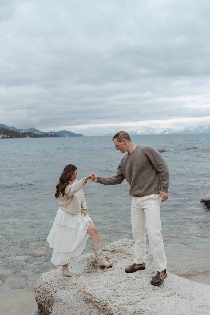 Man helping fiancé up large rock by holding her hand on a sandy beach in Lake Tahoe with storm clouds in background
