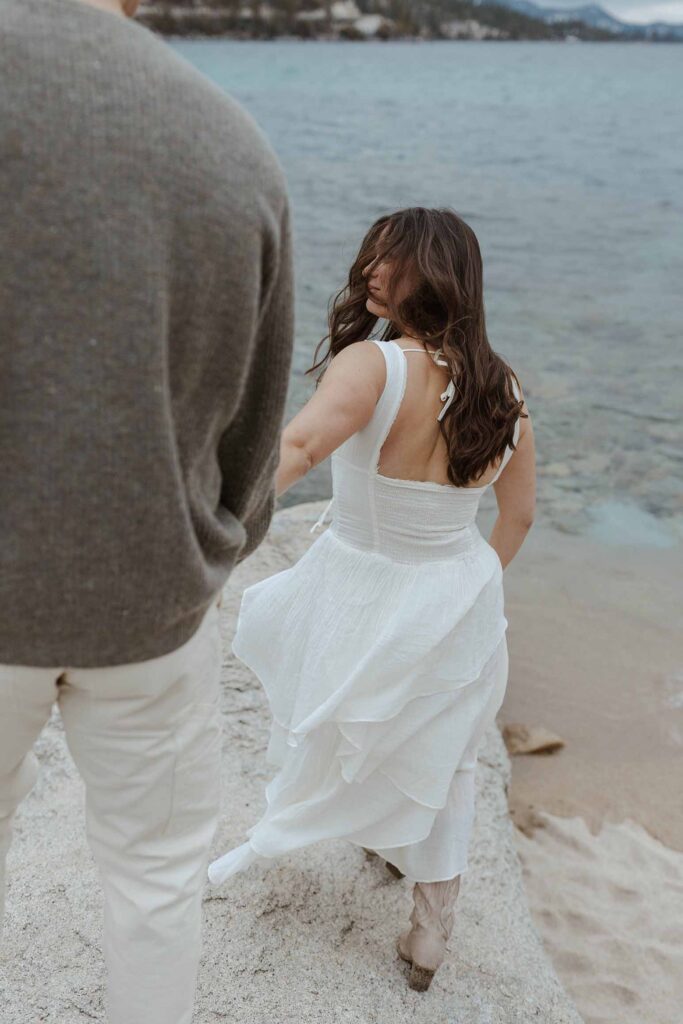 Woman leading fiancé while walking and looking back over her shoulder on a sandy beach in Lake Tahoe