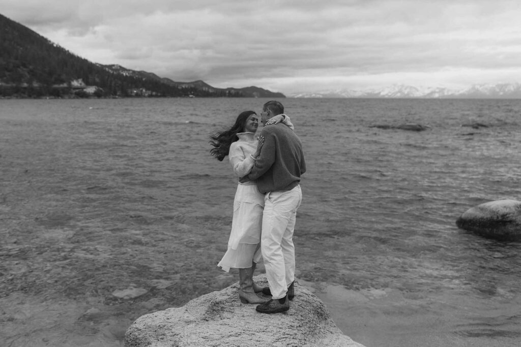 Engagement couple holding each other while standing on large rock surrounded by water on a beach in Lake Tahoe with storm clouds in background
