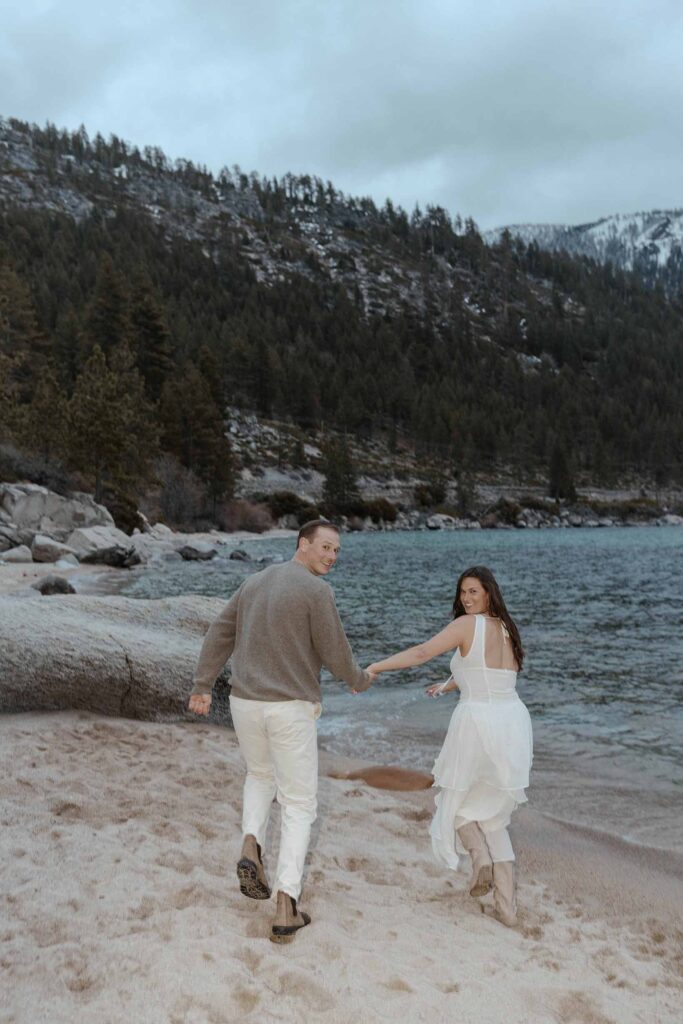Engagement couple holding hands while running along sandy beach in Lake Tahoe while looking back at camera with trees and storm clouds in background