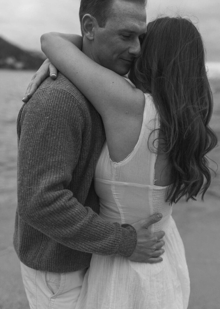 Engagement couple hugging each other while man has eyes closed on a sandy beach in Lake Tahoe with water in background