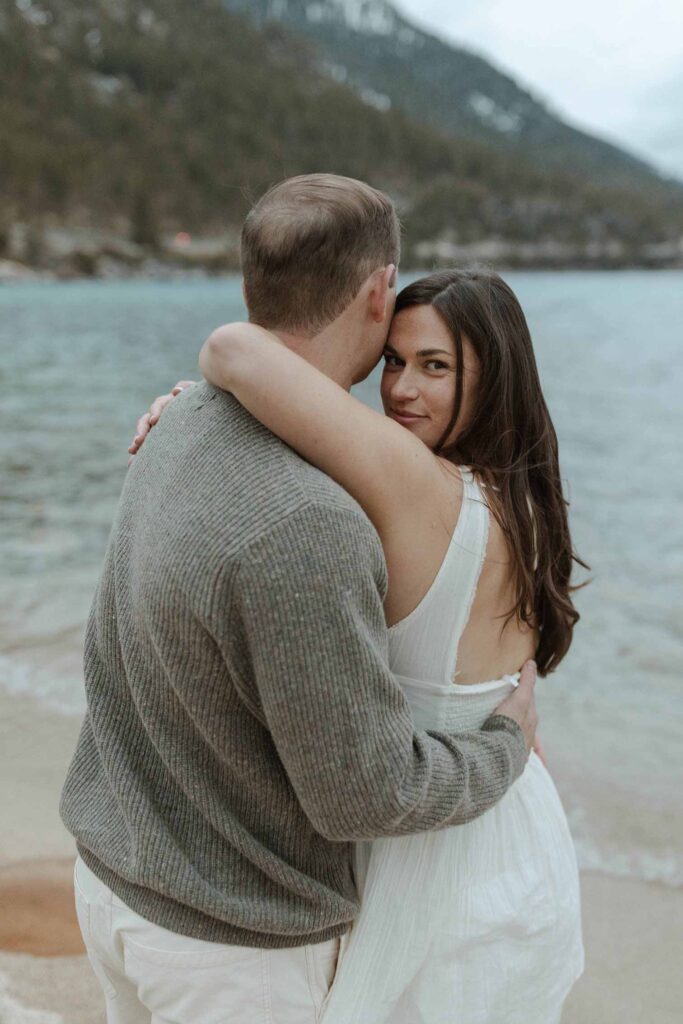 Engagement couple hugging while woman looks back at camera on a beach in Lake Tahoe with lake in background