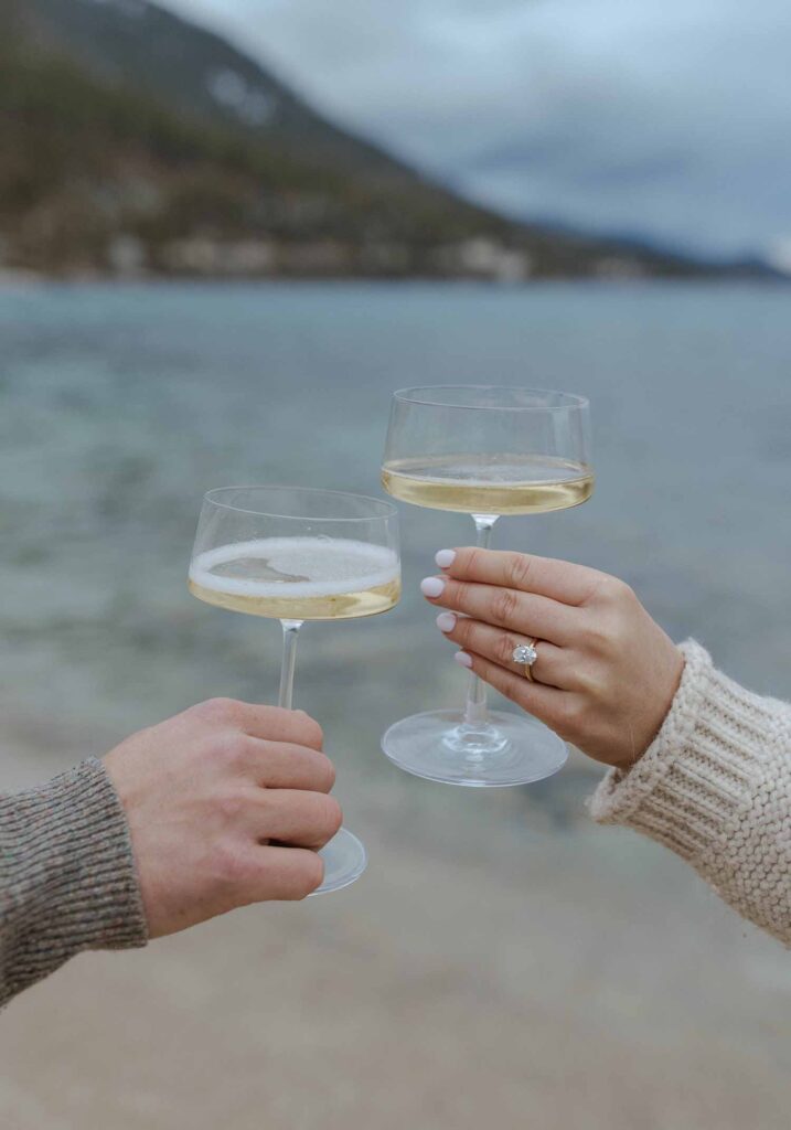 Couple toasting champagne together on a beach in Lake Tahoe while woman has engagement ring on finger