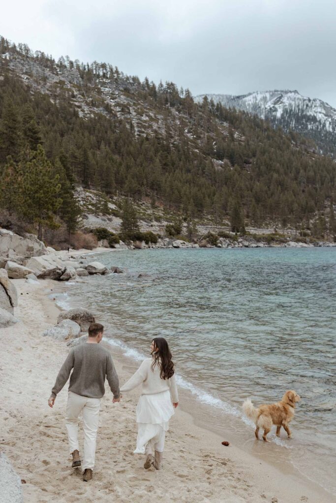 Engagement couple holding hands while walking along sandy beach in Lake Tahoe with dog and storm clouds in background