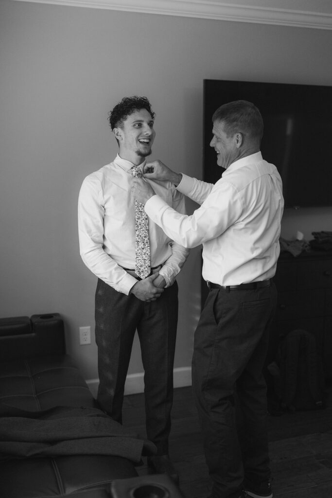 Groom's dad helping groom put on tie before elopement in Emerald Bay