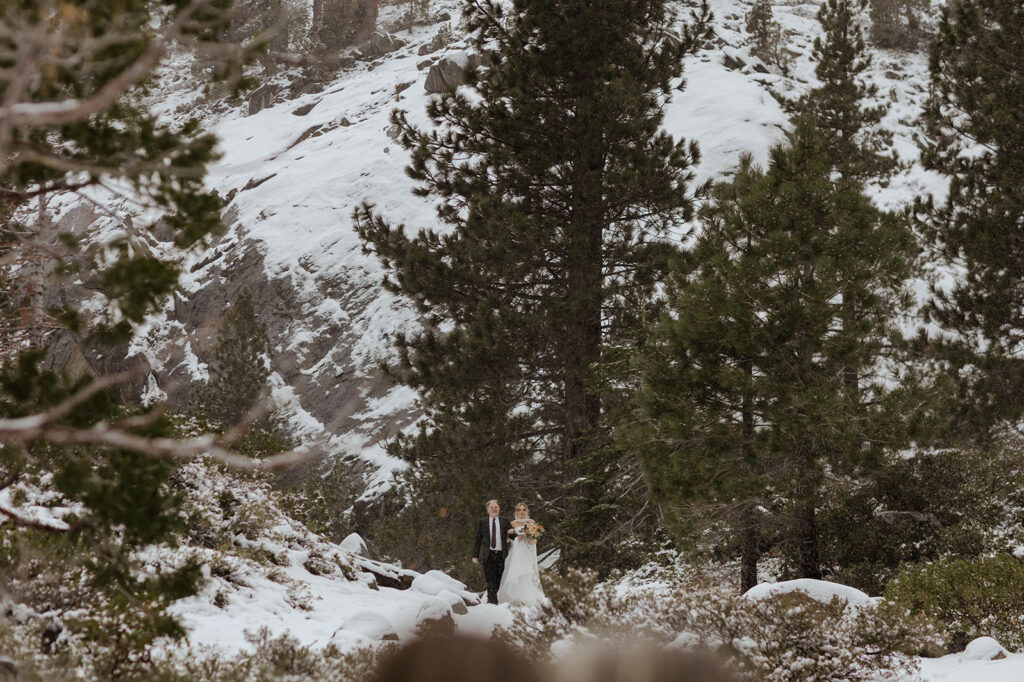 Bride walking down snowy mountain with dad to elopement ceremony in Emerald Bay with pine trees in background