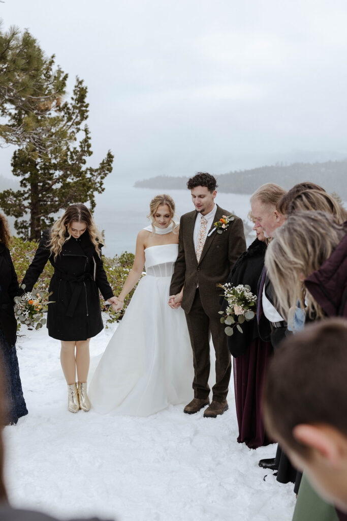 Elopement couple holding hands while praying with guests during snowy ceremony in Emerald Bay