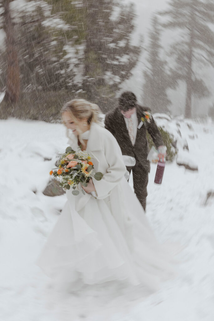 Elopement couple walking through snow together while holding flowers and wine bottles in Emerald Bay
