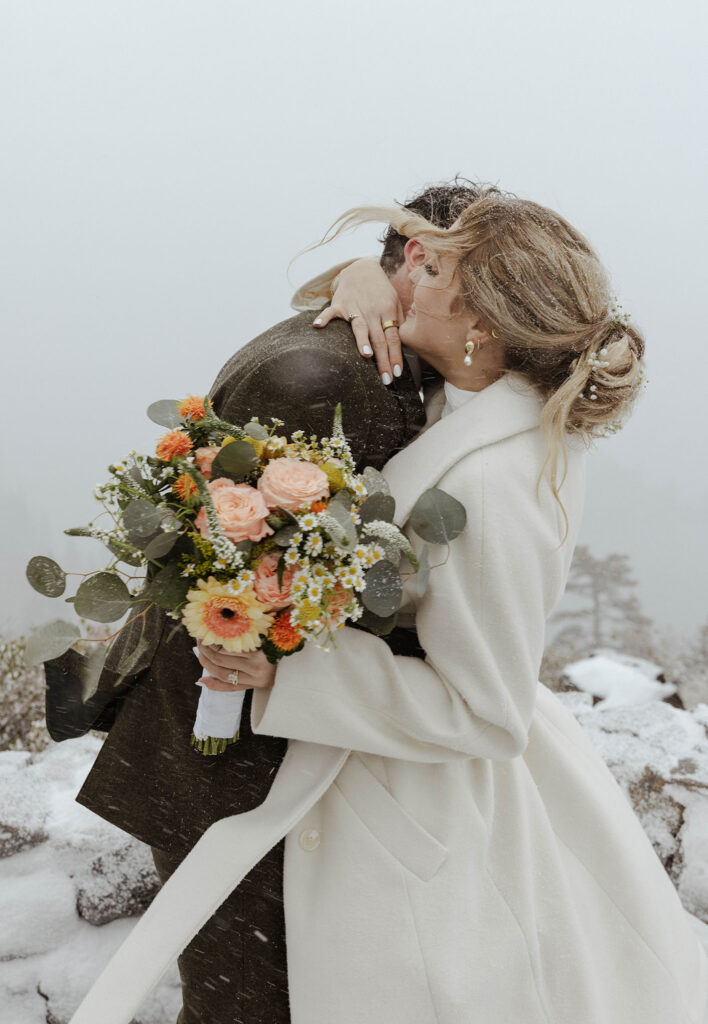 Elopement couple hugging while bride holds floral bouquet while snowing in Emerald Bay