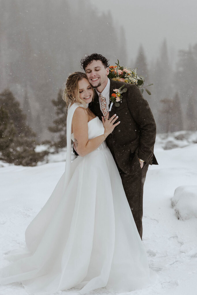Elopement couple smiling while hugging each other in the snow with trees in background in Emerald Bay