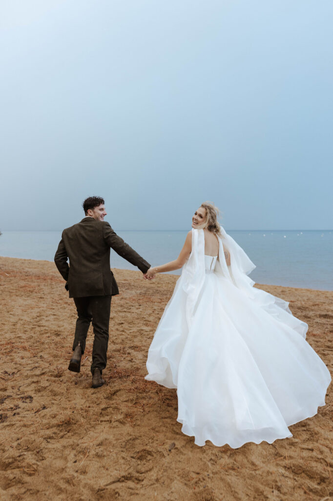 Elopement couple holding hands while running along sandy beach together and looking back at camera in Emerald Bay