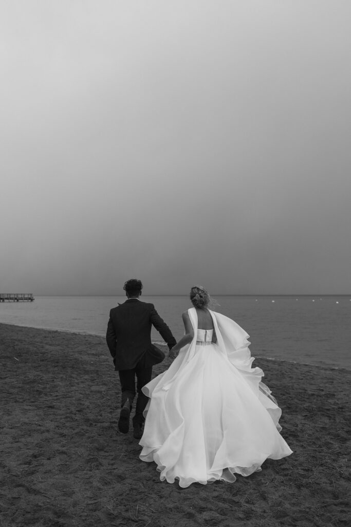 Elopement couple holding hands and running along sandy beach away from camera towards Lake Tahoe in Emerald Bay