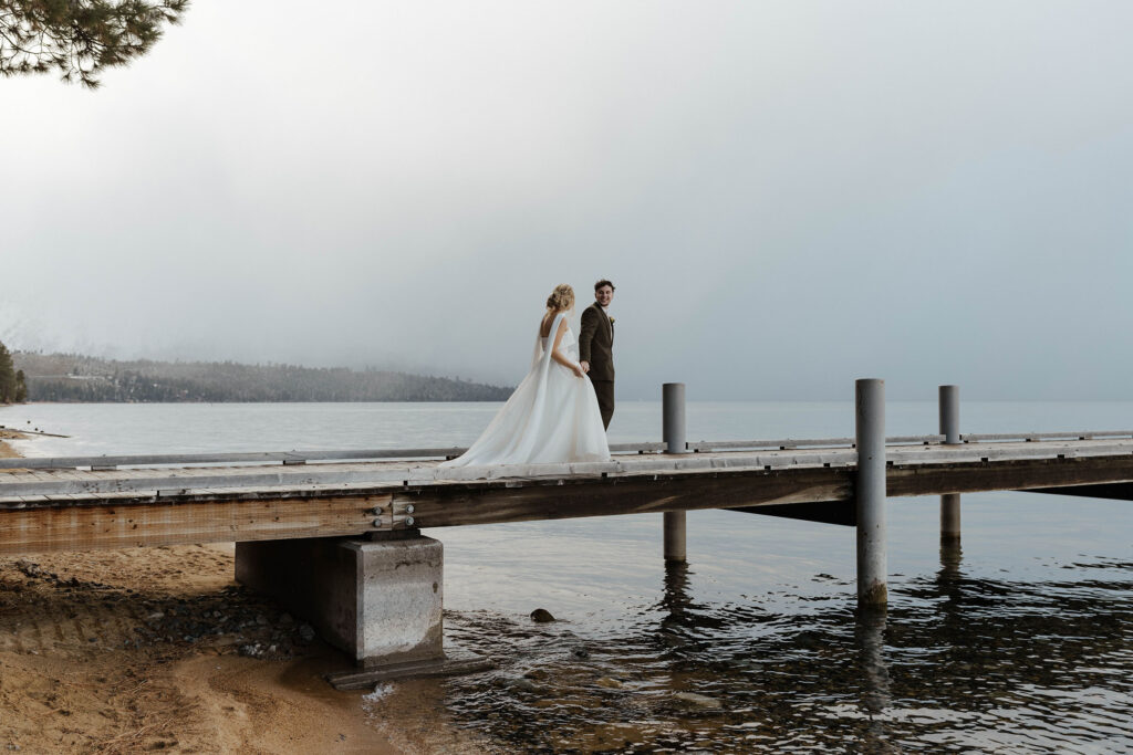 Elopement couple holding hands while walking along pier towards Lake Tahoe with clouds in background