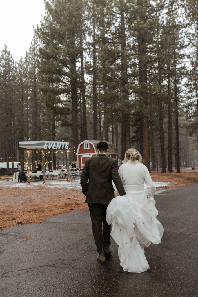 Groom holding brides dress while both walk toward elopement reception with tall trees in background
