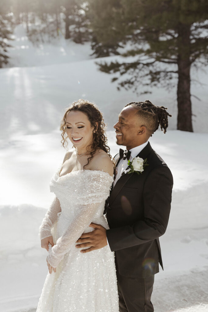 Groom holding wedding bride from behind while both smile and snow in the background