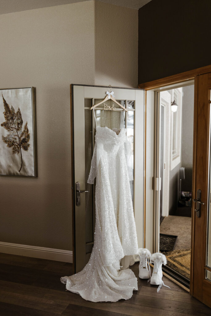 Wedding dress on wooden hanger in doorway next to white wedding boots for winter wedding at the Tannenbaum