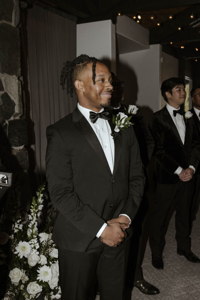 Groom smiling while standing at wedding ceremony watching bride walk down the aisle at the Tannenbaum with white floral bouquets behind him