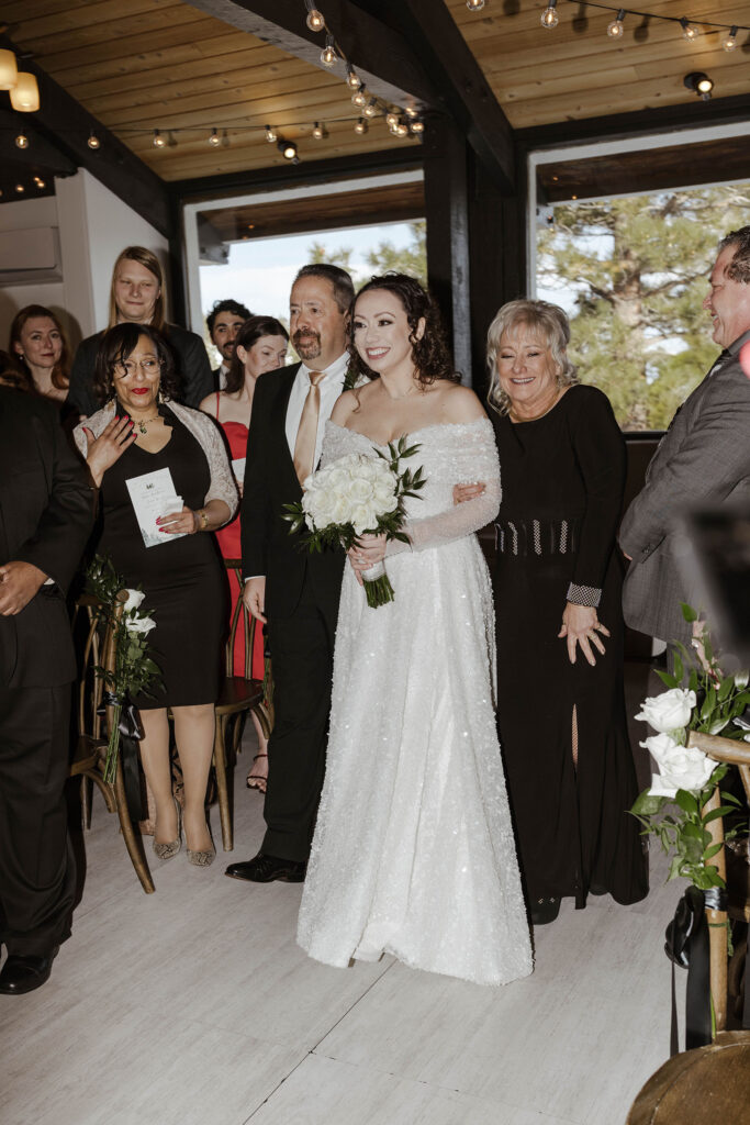 Bride smiling while walking down aisle at wedding ceremony with parents on either side and guests watching at the Tannenbaum
