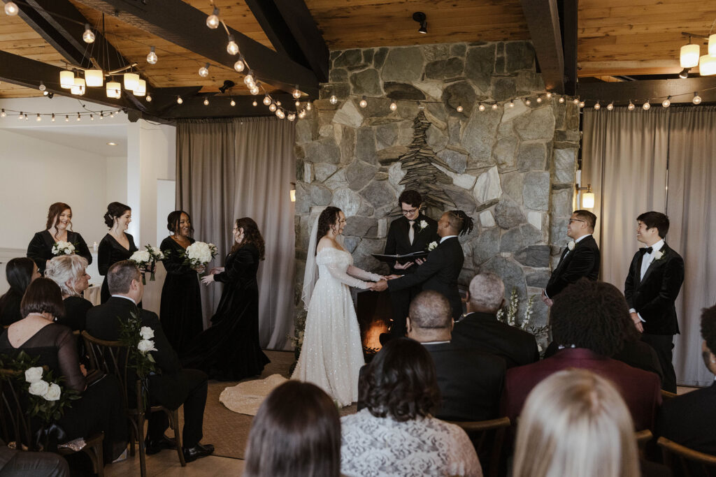 Wedding couple holding hands in front of stone fireplace during ceremony at the Tannenbaum with string lights above them