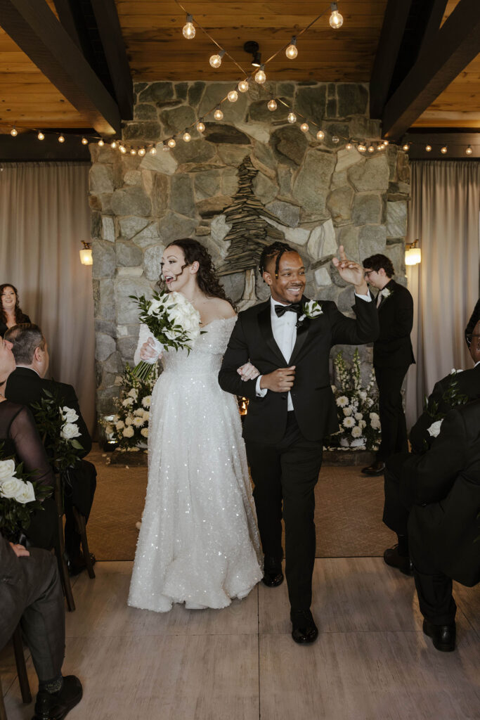 Wedding couple holding hands while walking down aisle and celebrating after ceremony at the Tannenbaum with stone fireplace and string lights behind them