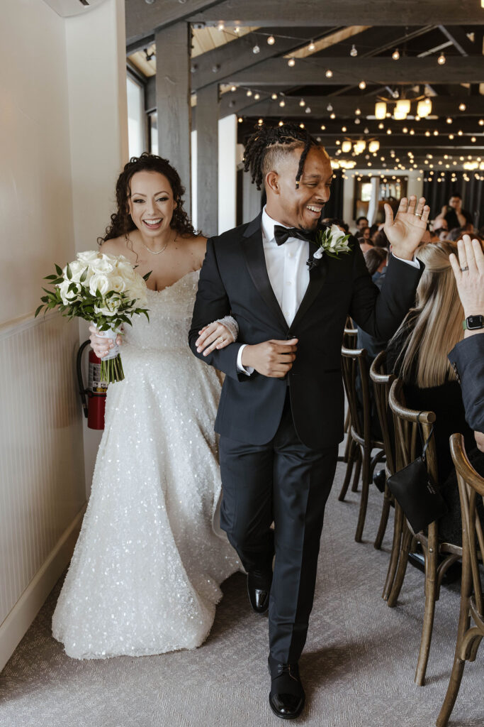 Wedding couple smiling while walking out of ceremony and waving to guests at the Tannenbaum with string lights in rafters behind them