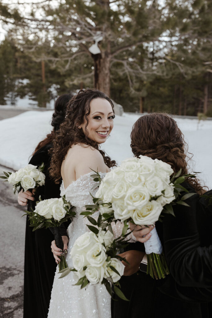 Wedding bride looking over shoulder at camera while smiling and standing next to bridesmaids with snow in background at the Tannenbaum