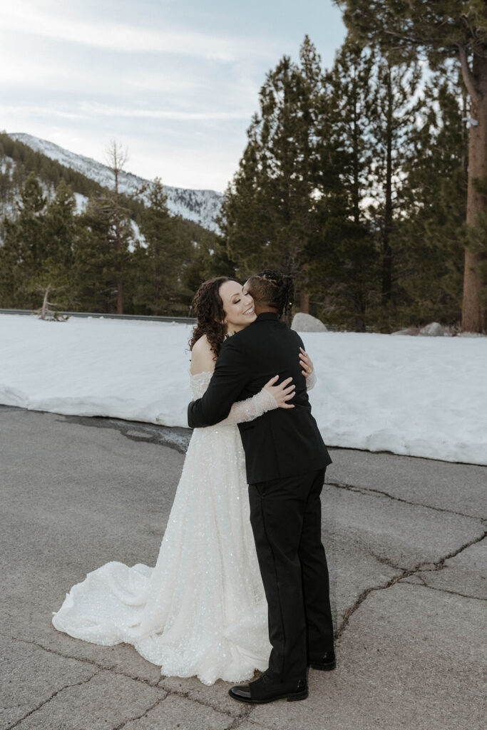 Wedding couple hugging in front of snow bank and pine trees while bride smiles