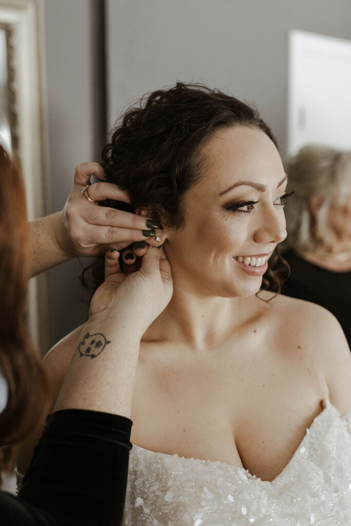 Bride's mom putting on wedding bride's earrings while she smiles inside at the Tannenbaum