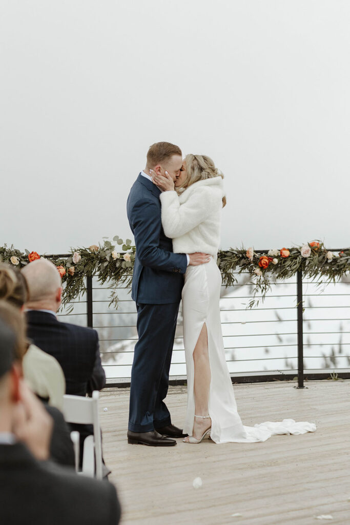 Wedding couple first kiss during ceremony at High camp at Palisades Tahoe with floral decorations on railing 