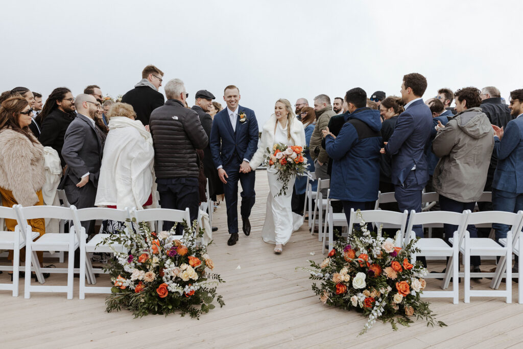Wedding couple smiling and holding hands walking out of ceremony at Palisades Tahoe while guests stand and clap