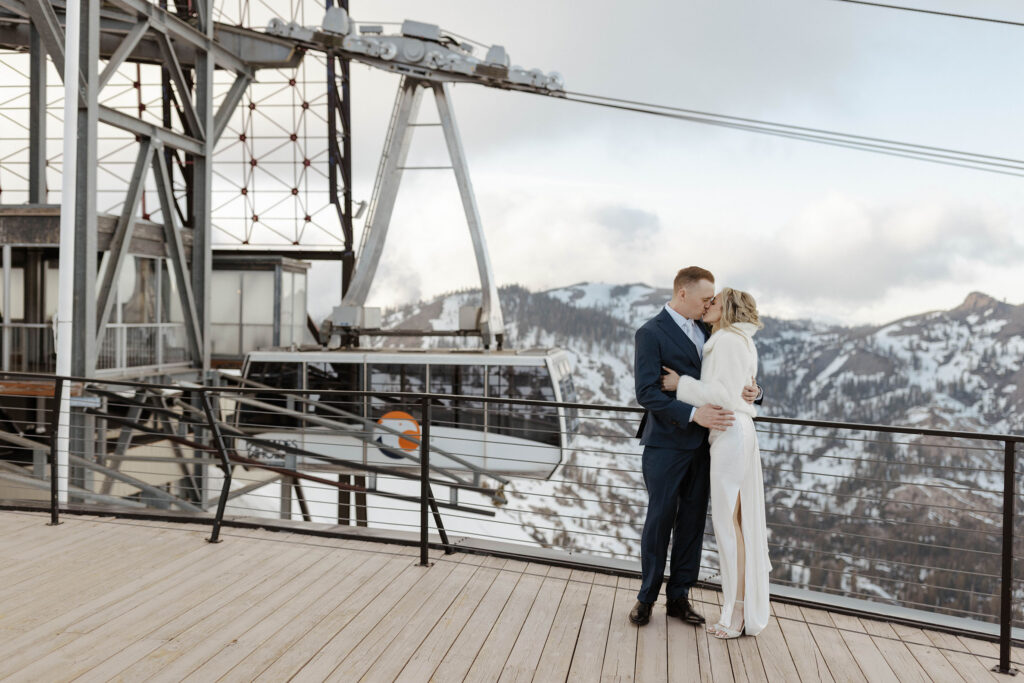 Wedding couple holding and kissing each other while standing on overlook at Palisades Tahoe with snow covered mountains in background