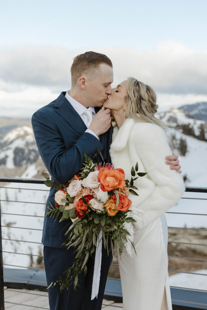 Wedding couple kissing while groom holds bride's chin and she holds floral bouquet at High camp at Palisades Tahoe with snowy mountains in background