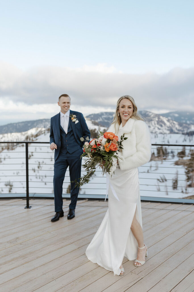 Bride smiling at camera while holding colorful floral bouquet and groom smiles at her from behind at High camp at Palisades Tahoe