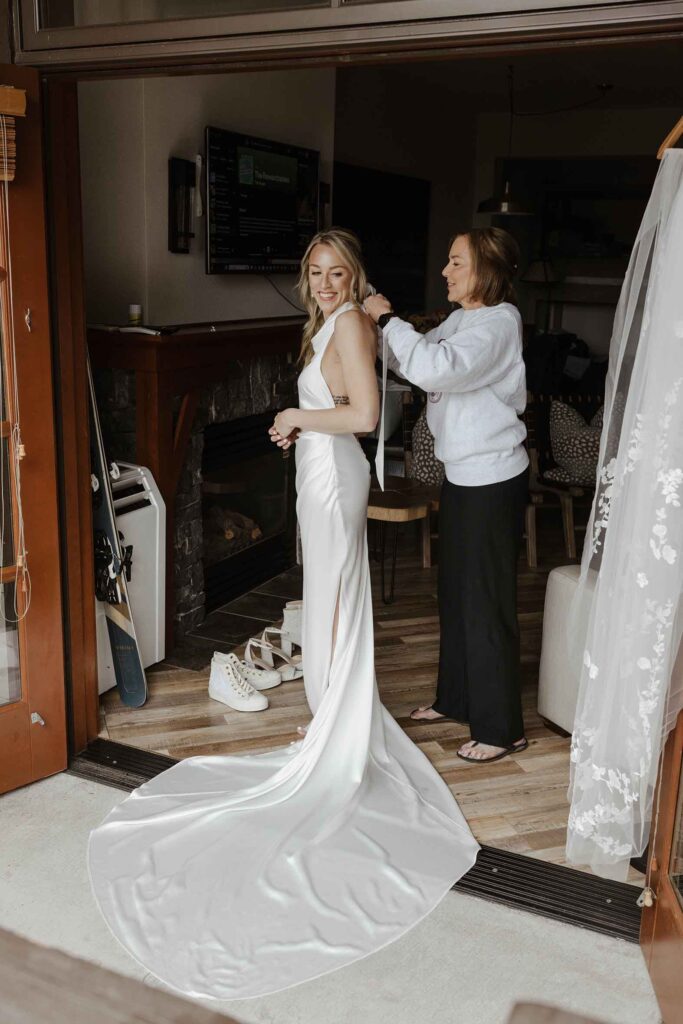 Bride looking over shoulder and smiling while mom ties her wedding dress at Palisades Tahoe