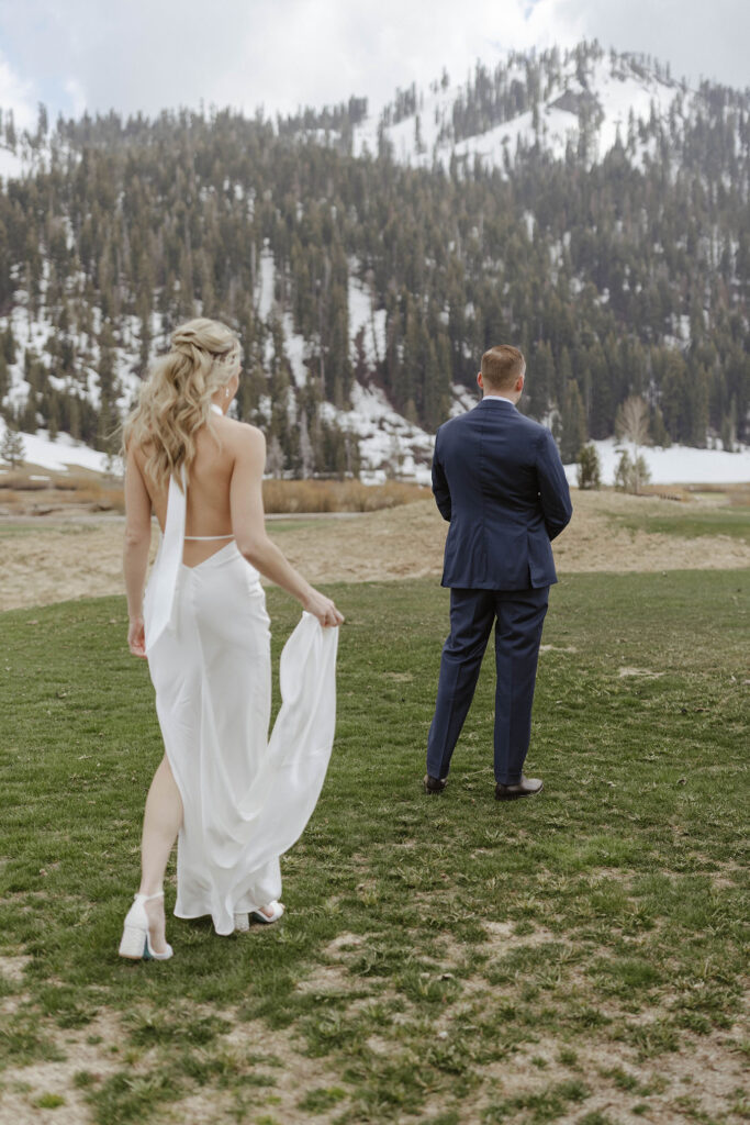 Bride holding wedding dress and walking up behind groom in grass field with snowy mountains in background