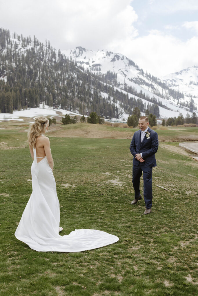 Groom excited after turning around and seeing bride in wedding dress during first look in grass field with snowy mountains in background
