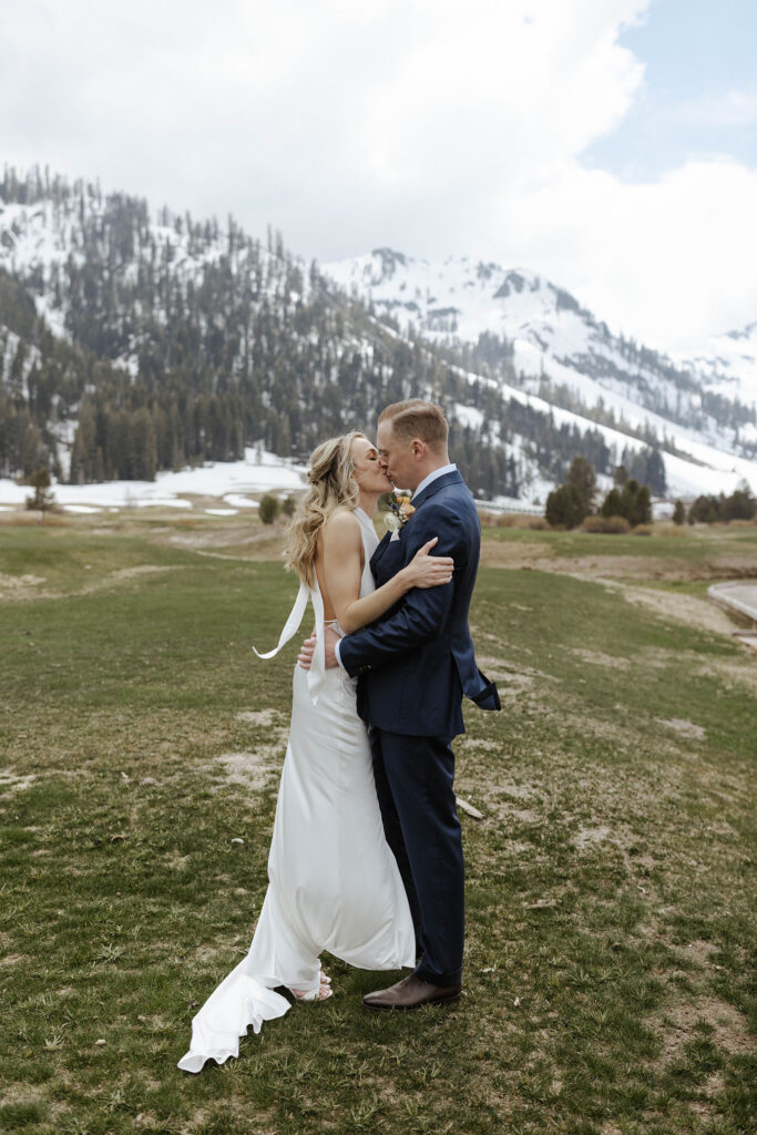 Wedding couple holding each other and kissing while in grass field with snowy mountains in background at Palisades Tahoe