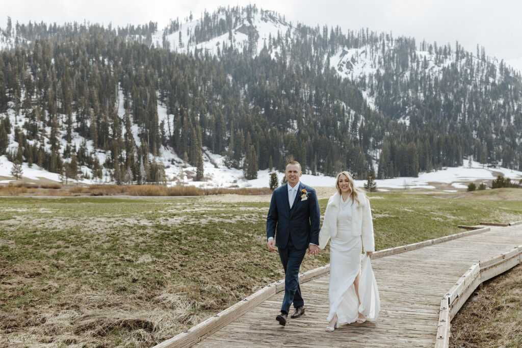 Wedding couple holding hands while walking along wooden walkway together towards camera with snowy mountains in background at Palisades Tahoe