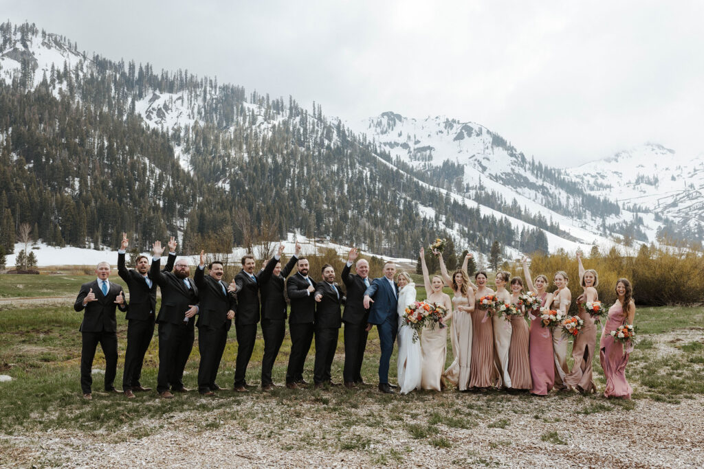 Entire bridal party celebrating while standing next to each other with wedding couple in the middle and snowy mountains in background at Palisades Tahoe