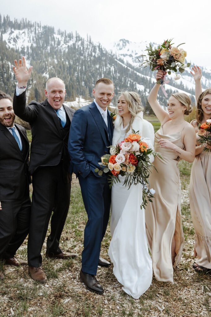 Wedding couple smiling while groomsmen and bridesmaids celebrate on either side of them with snowy mountains in background